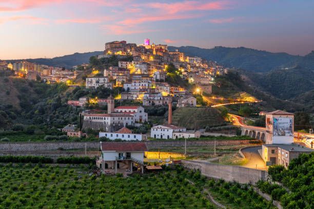 Illuminated hilltop town at dusk with surrounding greenery and structures.