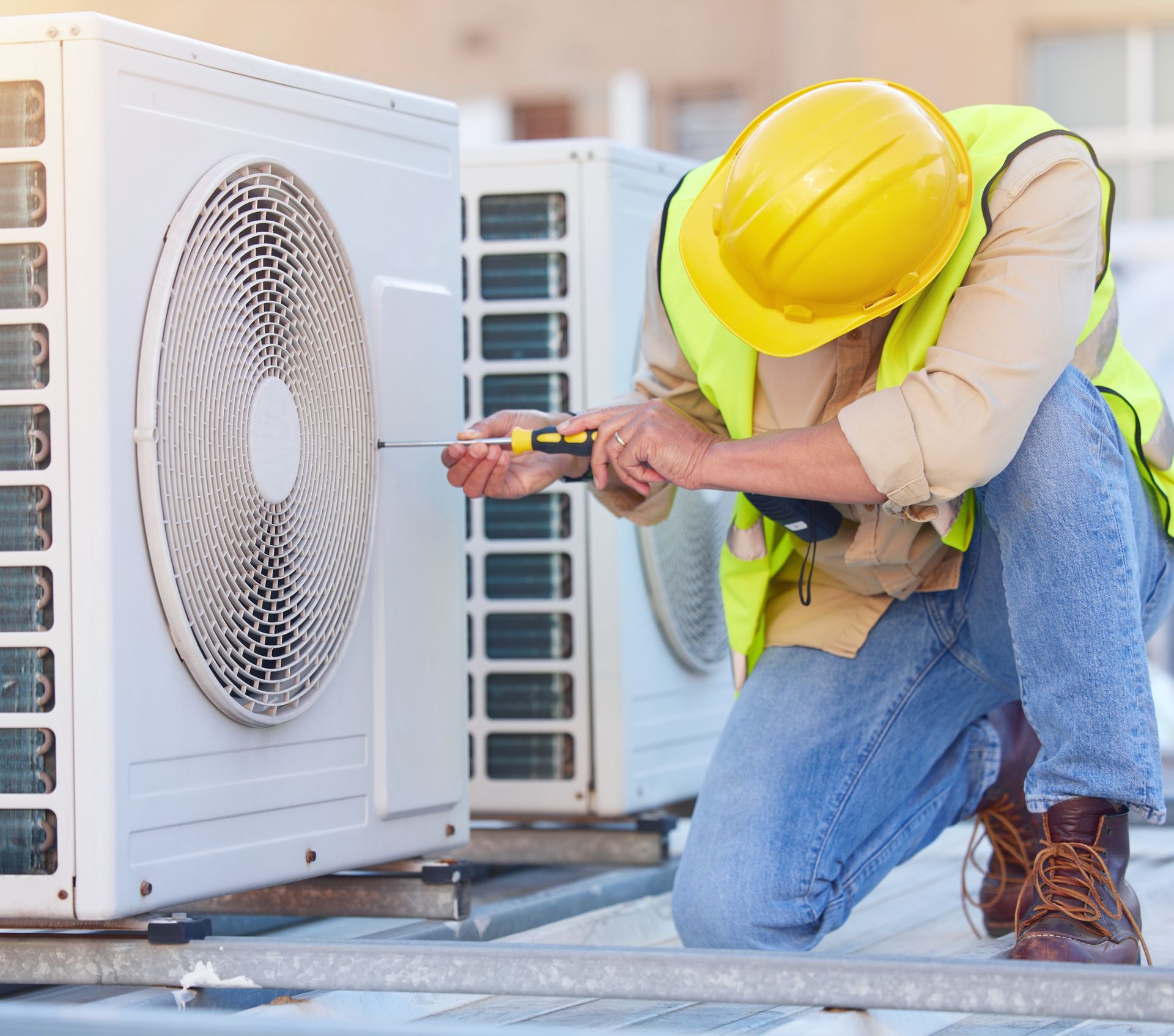 A male technician checks an AC unit fan with a screwdriver, on top of a roof.