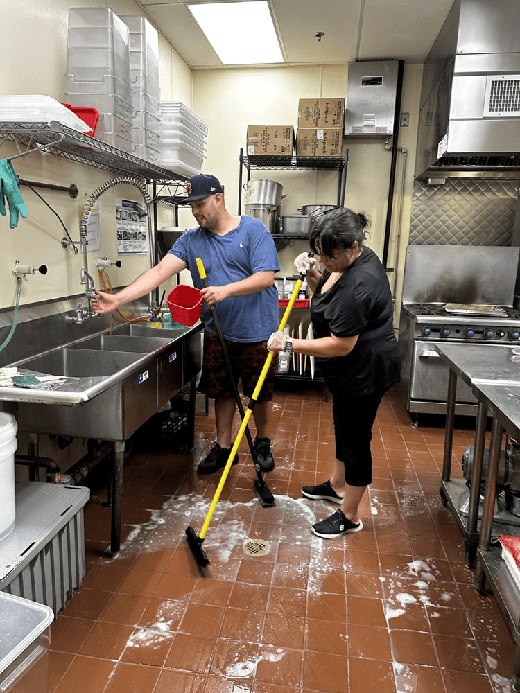 A man and a woman are cleaning a kitchen floor with a mop.