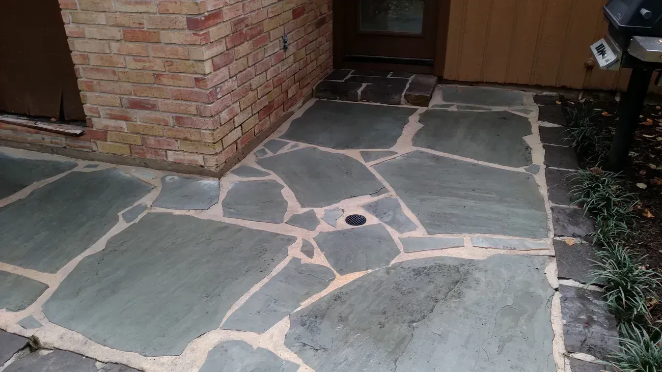 Flagstone patio with brick wall and wooden siding. Green-gray stones with light grout. Black edging and some greenery.
