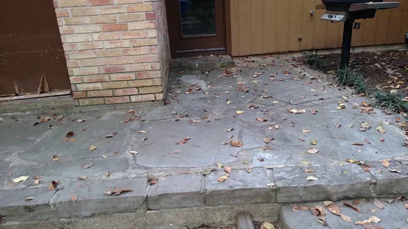 Concrete patio with scattered leaves, leading to a door next to a brick wall.