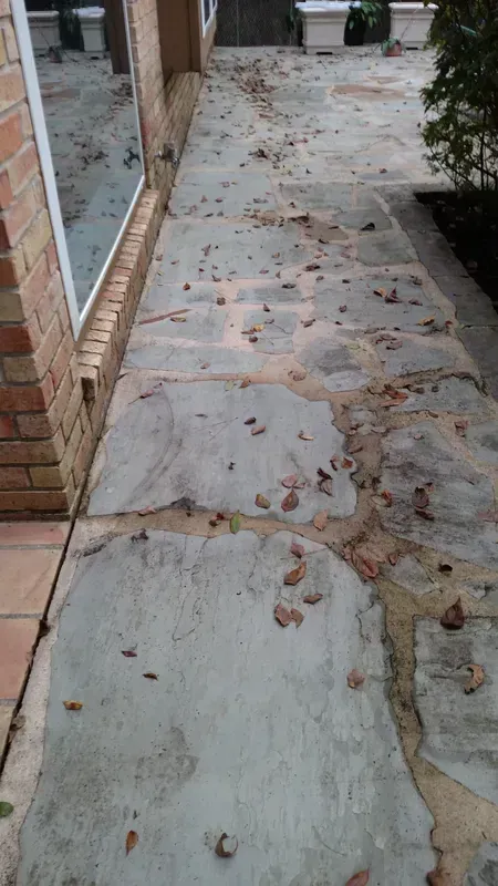 Concrete walkway with exposed aggregate, debris, next to a brick wall and window.