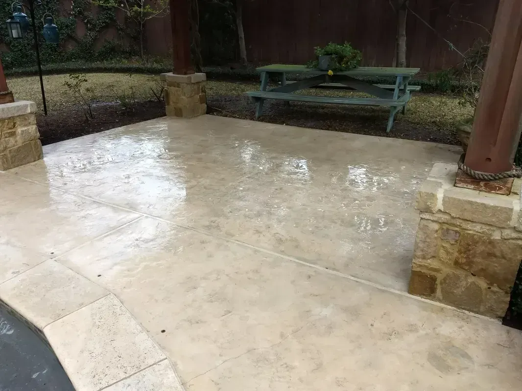 Wet concrete patio with limestone trim, beneath a pergola. A bench sits in the background.