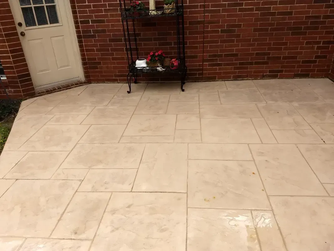 Tan stone patio against a brick wall, with a door on the left and a black plant stand in the center.