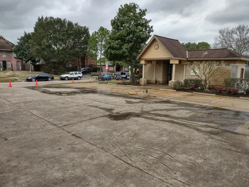 Paved area in front of a tan building. Several vehicles and trees are visible under an overcast sky.