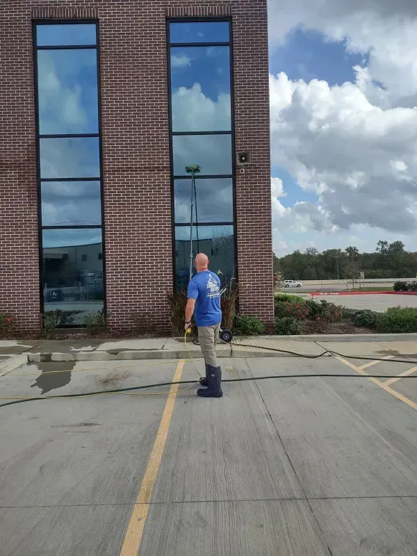 Man cleaning a tall window with a long-handled tool on a building exterior. Cloudy sky and parking lot.