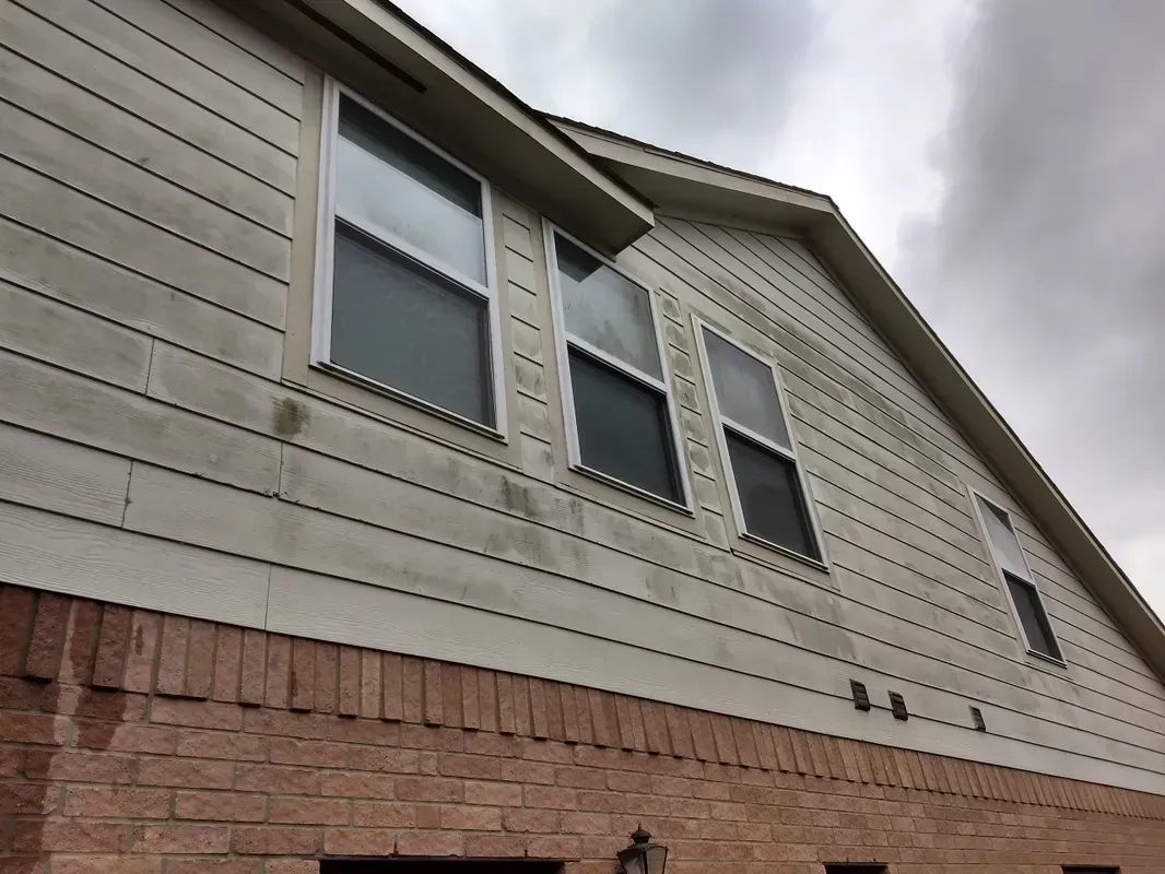 Exterior of a house with weathered siding. Brick foundation, windows, and a cloudy sky.