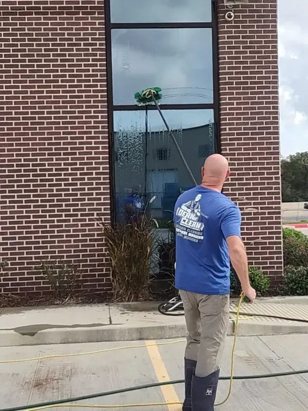 Man washing a tall window on a brick building using an extended brush, standing outside on a sunny day.