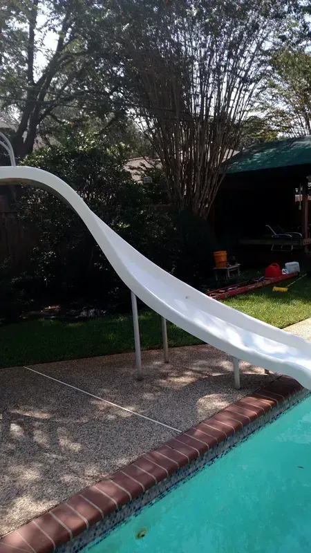 White pool slide next to a pool with a brick border. Green grass and foliage in the background.