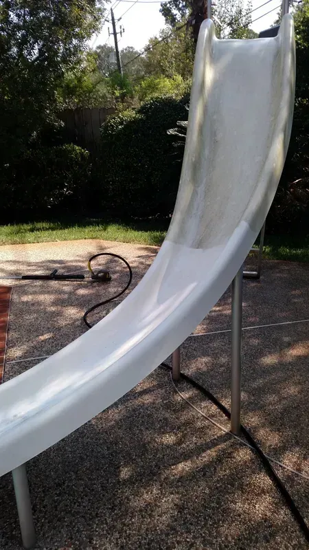White, curved slide on metal supports outdoors, with a dark hose and greenery in background.
