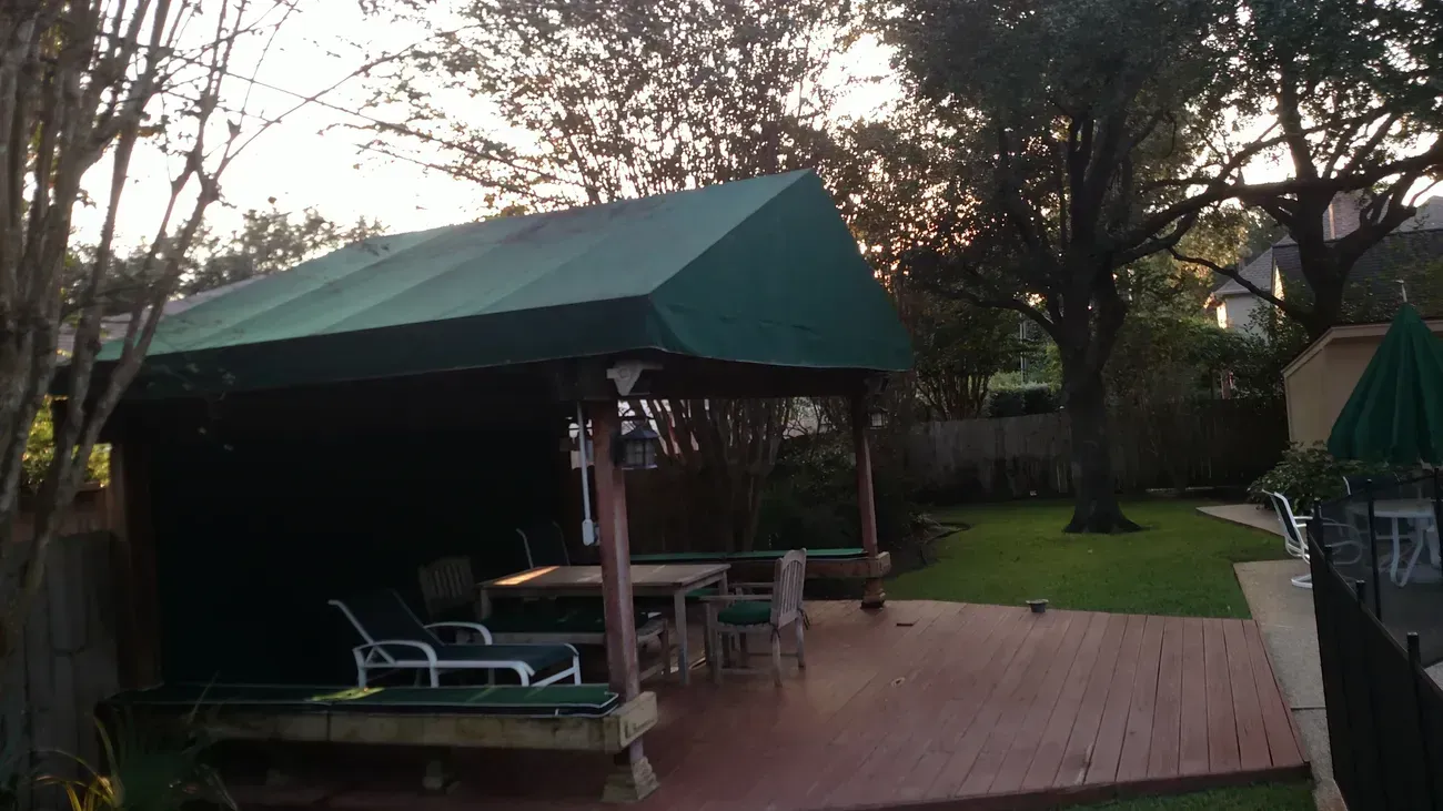 Green-roofed backyard pavilion over deck with outdoor furniture; lawn and trees in background.