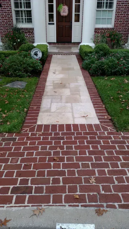 Brick walkway leading to a brown door with white columns. The path is lined with green shrubs and grass.