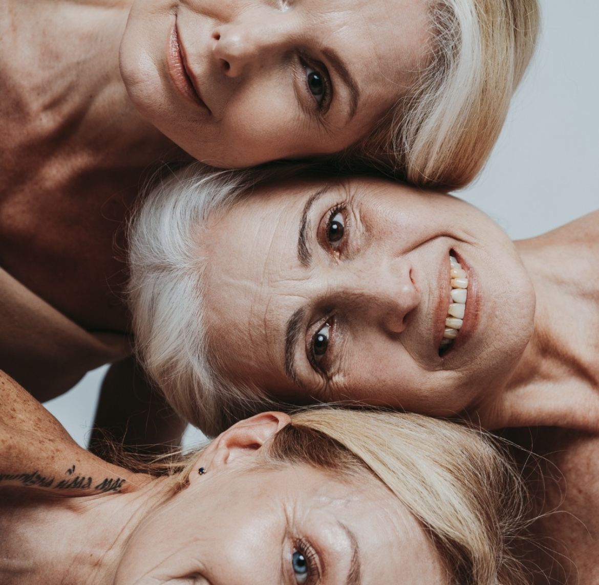 Three women with light hair smiling, bodies close together, against a white backdrop.