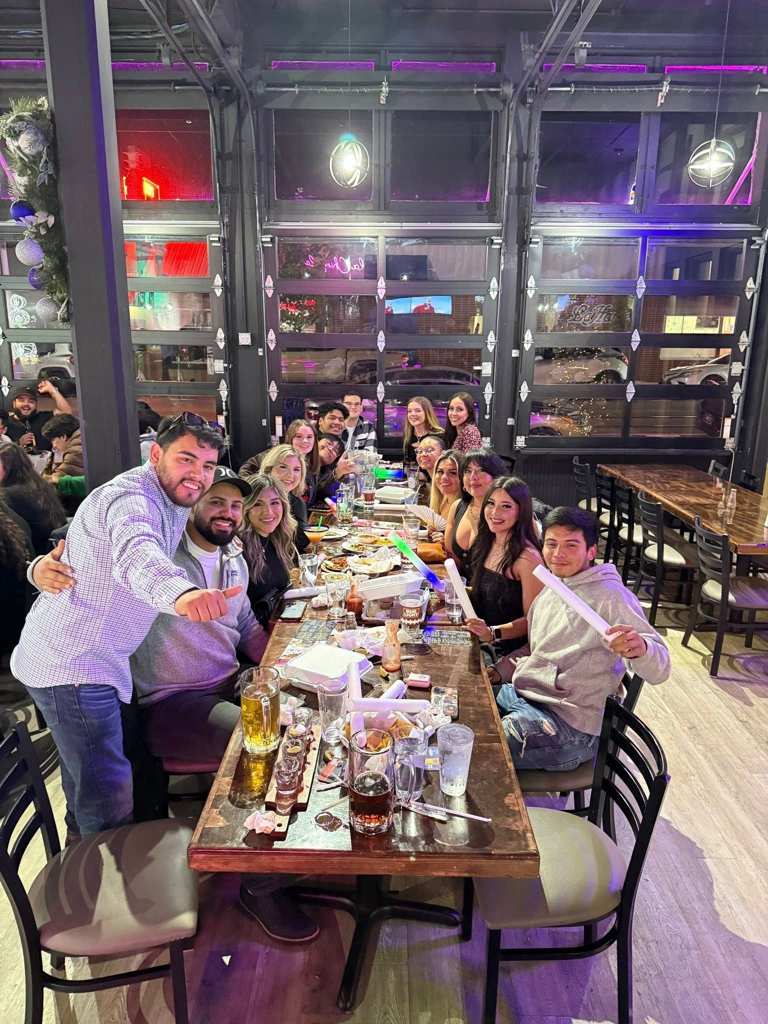 Group dining at a long restaurant table, with people smiling and raising drinks in a festive indoor setting Group dining at a long restaurant table, with people smiling and raising drinks in a festive indoor setting