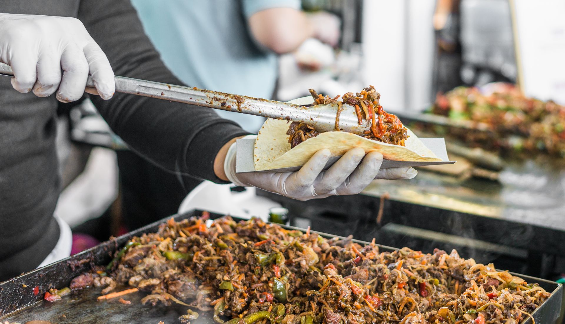 Gloved hands plating cooked food from a street food grill into a paper tray