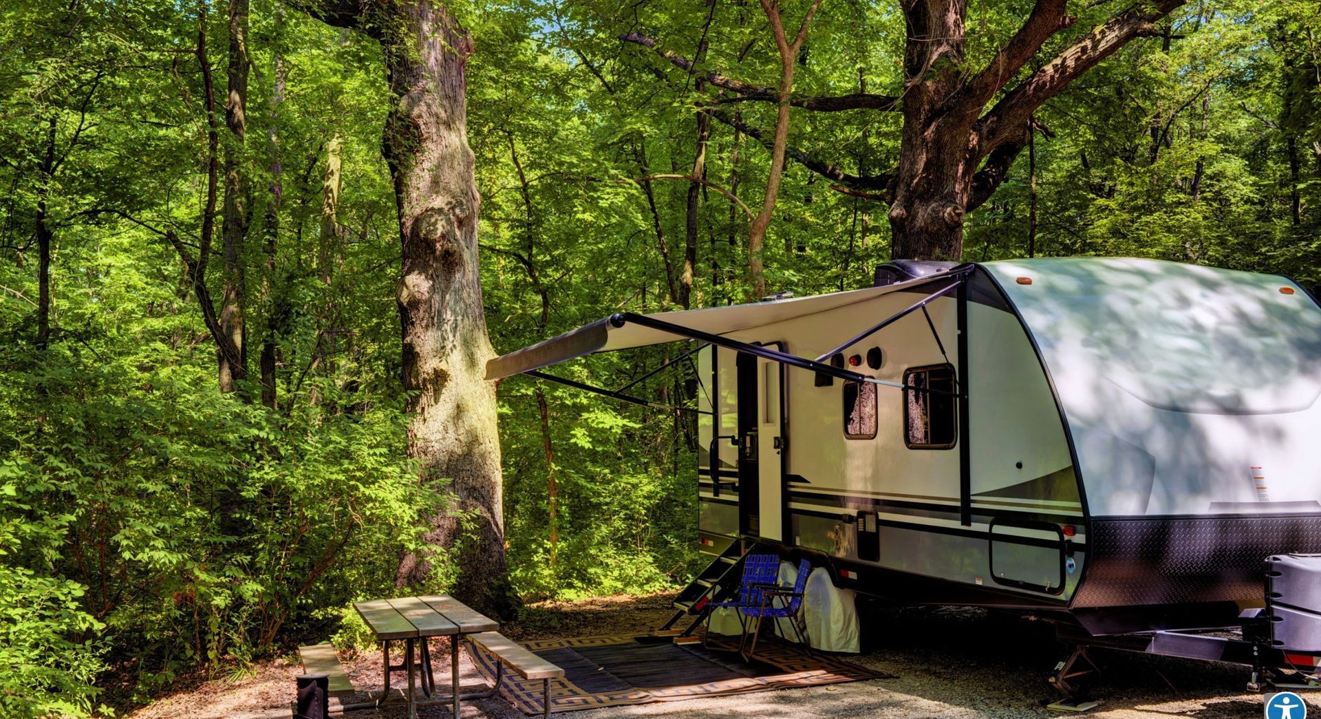 RV parked in a wooded campsite with awning extended. Table and trees in view.