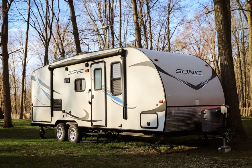Travel trailer parked in a wooded area, trees in the background, awning extended, gray and white exterior.