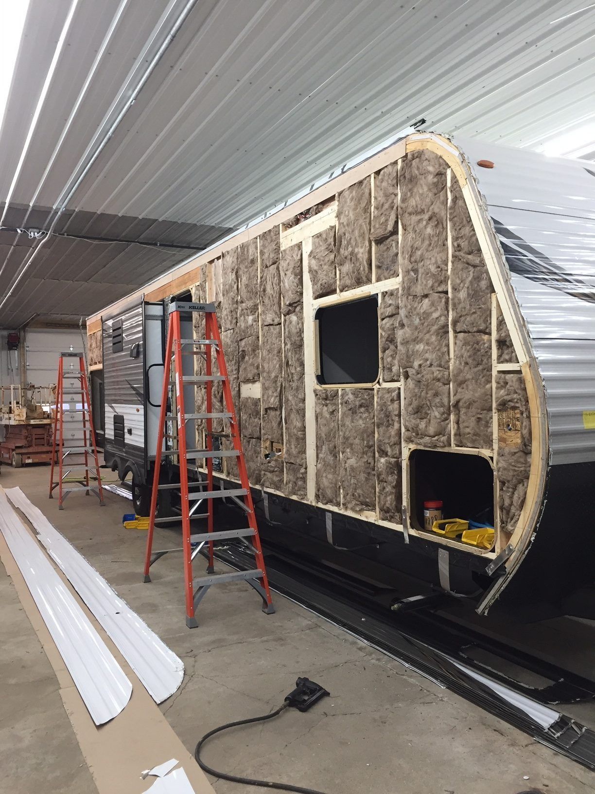 RV exterior under construction; insulation exposed. Ladders, tools, and metal siding present in a workshop setting.