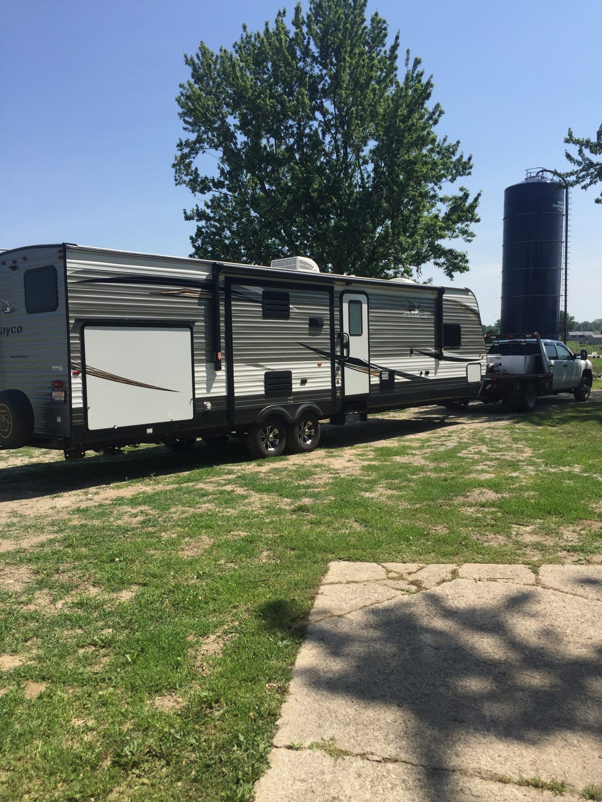 A travel trailer hitched to a truck on grass, with a tree and silo in the background.