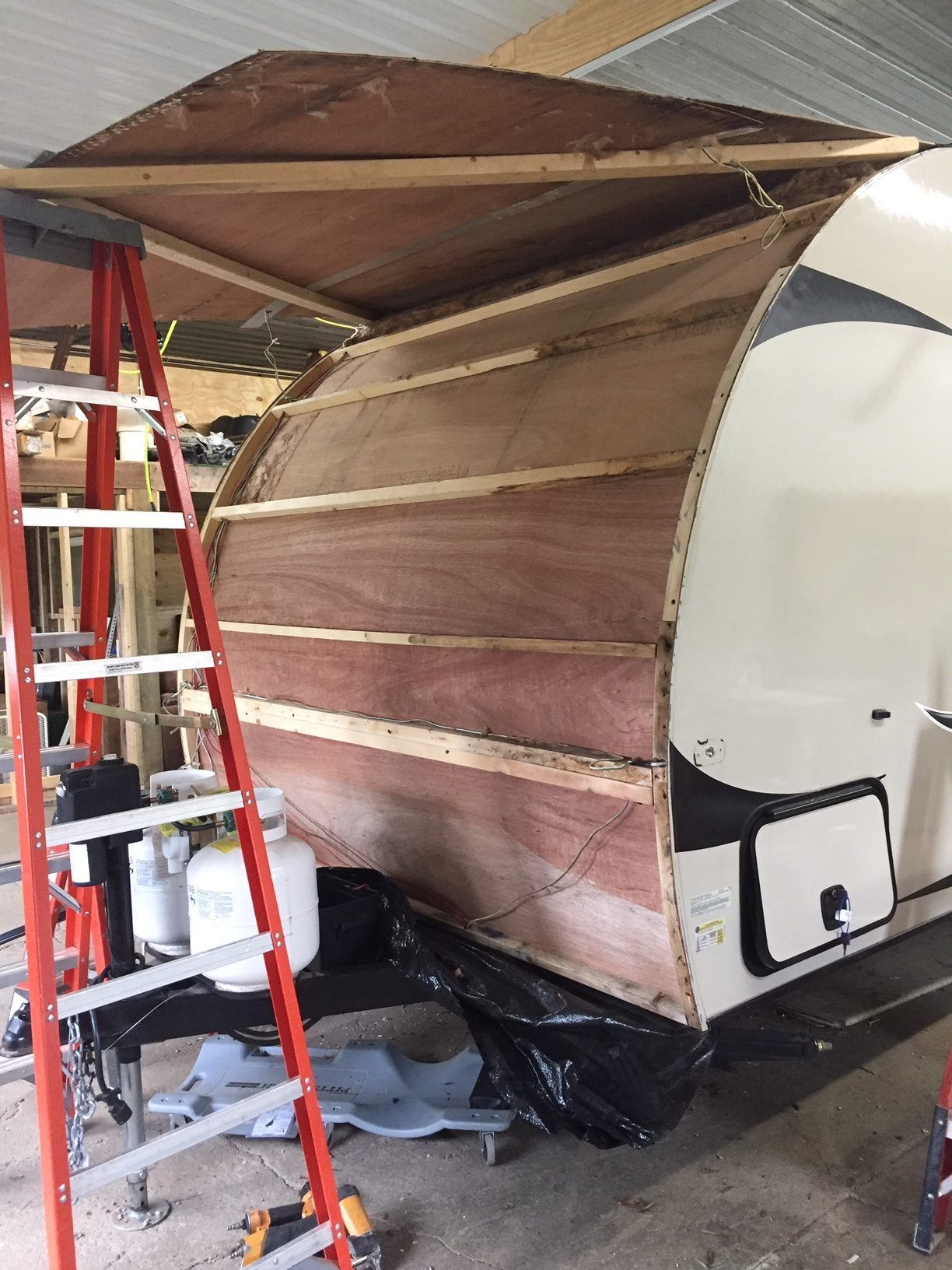 RV side under construction, with wooden frame and plywood. A-frame ladder visible.
