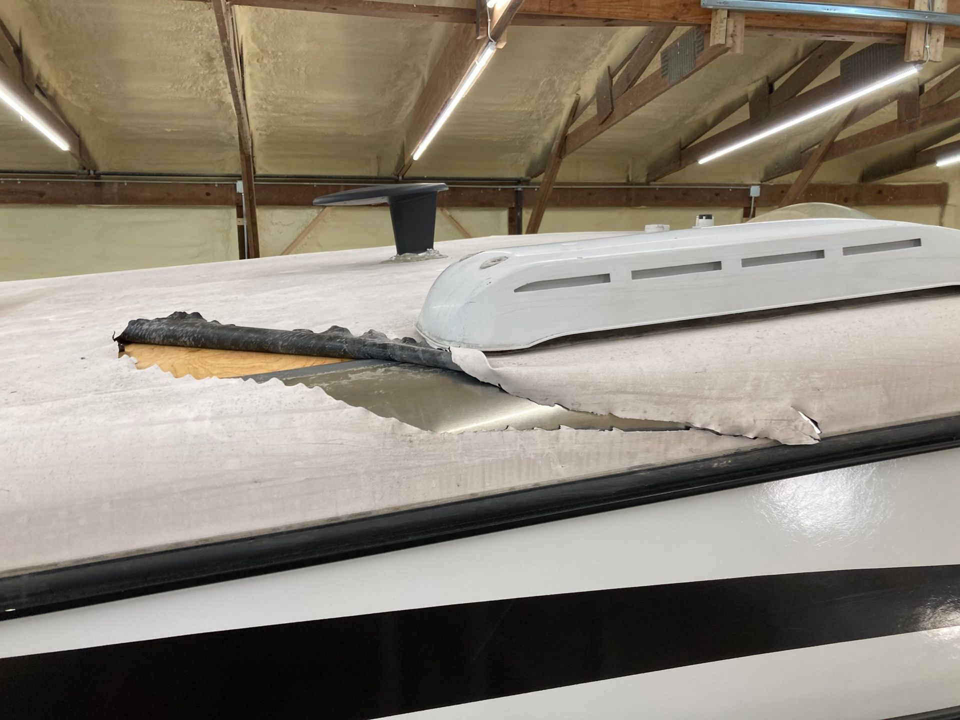 Damaged RV roof with torn white covering, exposing underlying wood. Black trim, air vent, and AC unit visible.