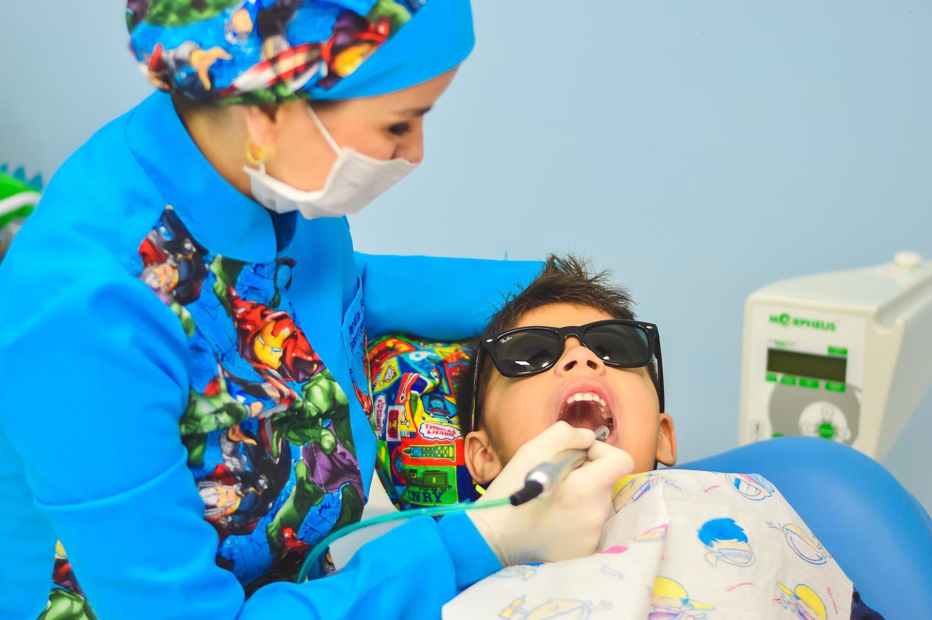 Dentist in blue scrubs examines a child's teeth with dental tools, child wears sunglasses, dental setting.
