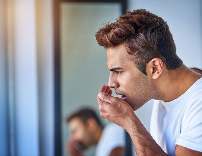 A man is covering his mouth while looking at himself in the mirror.