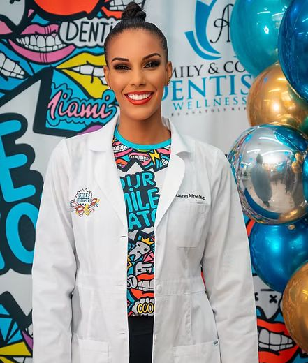 Woman in lab coat smiles, stands near balloons and a graffiti wall.
