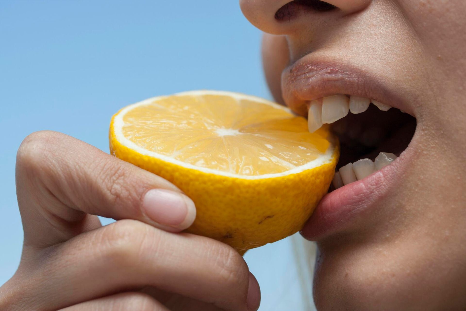 Person biting into a halved yellow lemon against a blue sky.