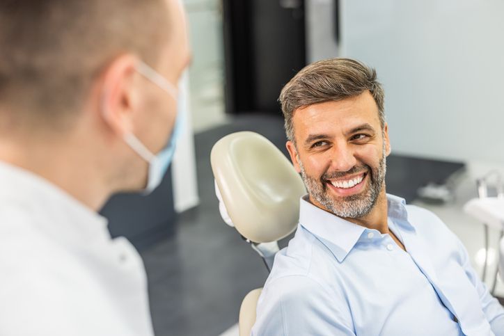 Man smiles at dentist in a dental chair. Dentist wears a mask, pale-toned room.