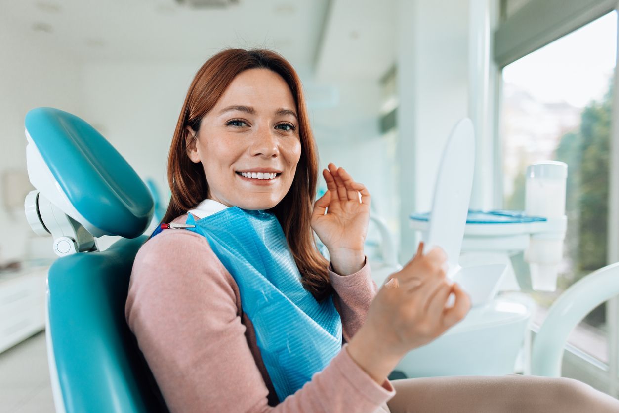 Woman in a dentist chair looking at her teeth in a mirror, smiling. Blue bib, white dental office setting.