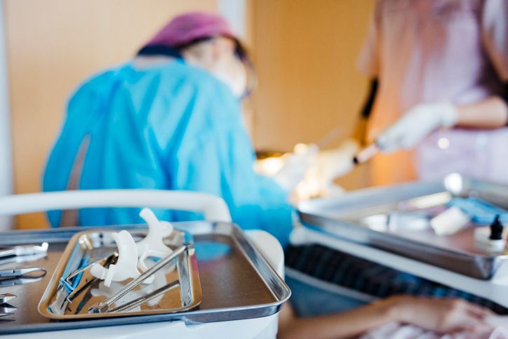 Surgical tools on tray in operating room; surgeon in blue scrubs operating. Assistant with needle.