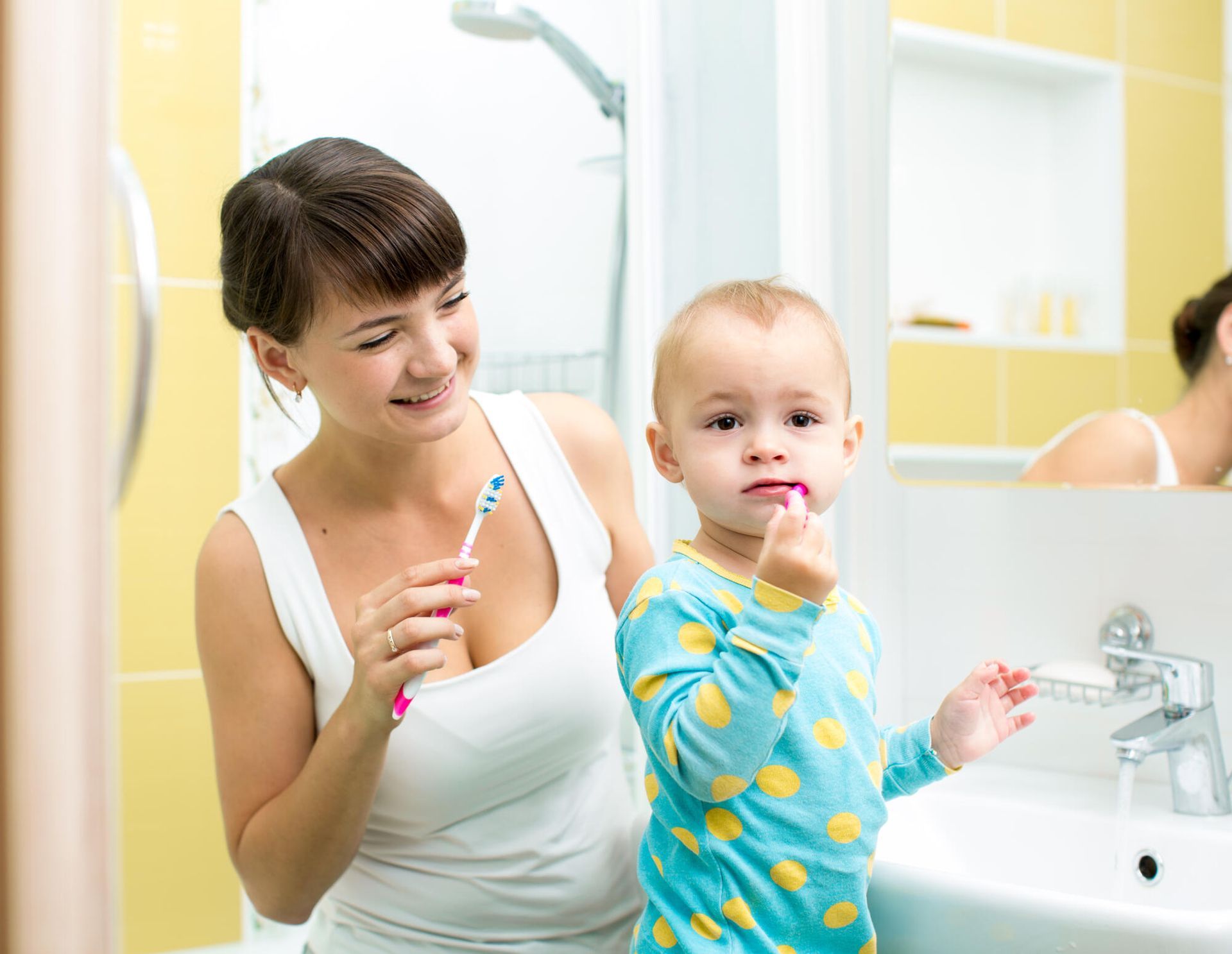 A woman and a young child in a yellow-tiled bathroom brushing their teeth together in front of a mirror.