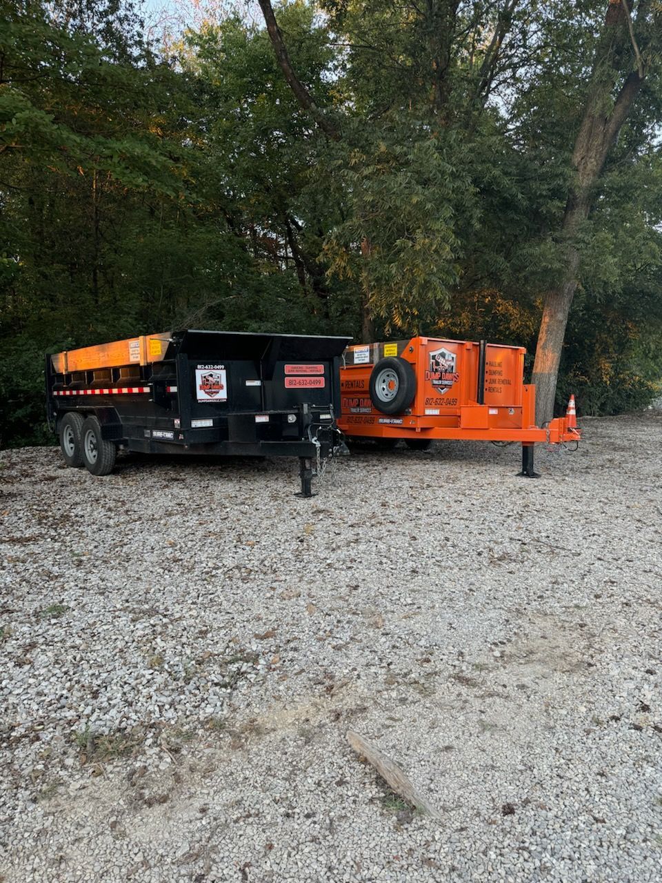 A dumpster and a trailer are parked in a gravel lot.