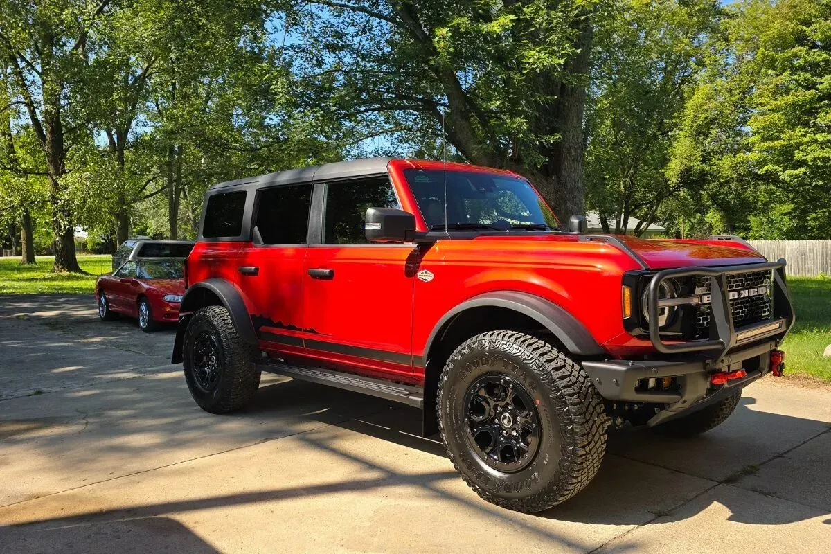 Red Ford Bronco with black accents parked on pavement. Trees and a car in the background.