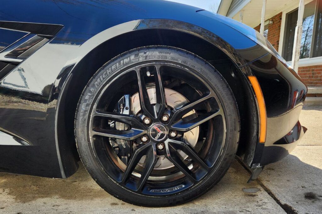 Black sports car's front wheel, showing a black rim and tire. Car parked on a sunny day.