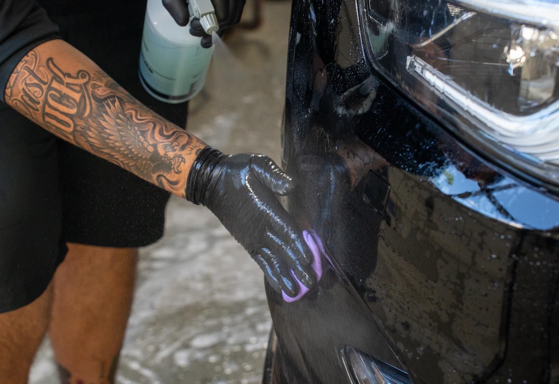 Person in black gloves spraying and wiping a black car, revealing a tattoo.
