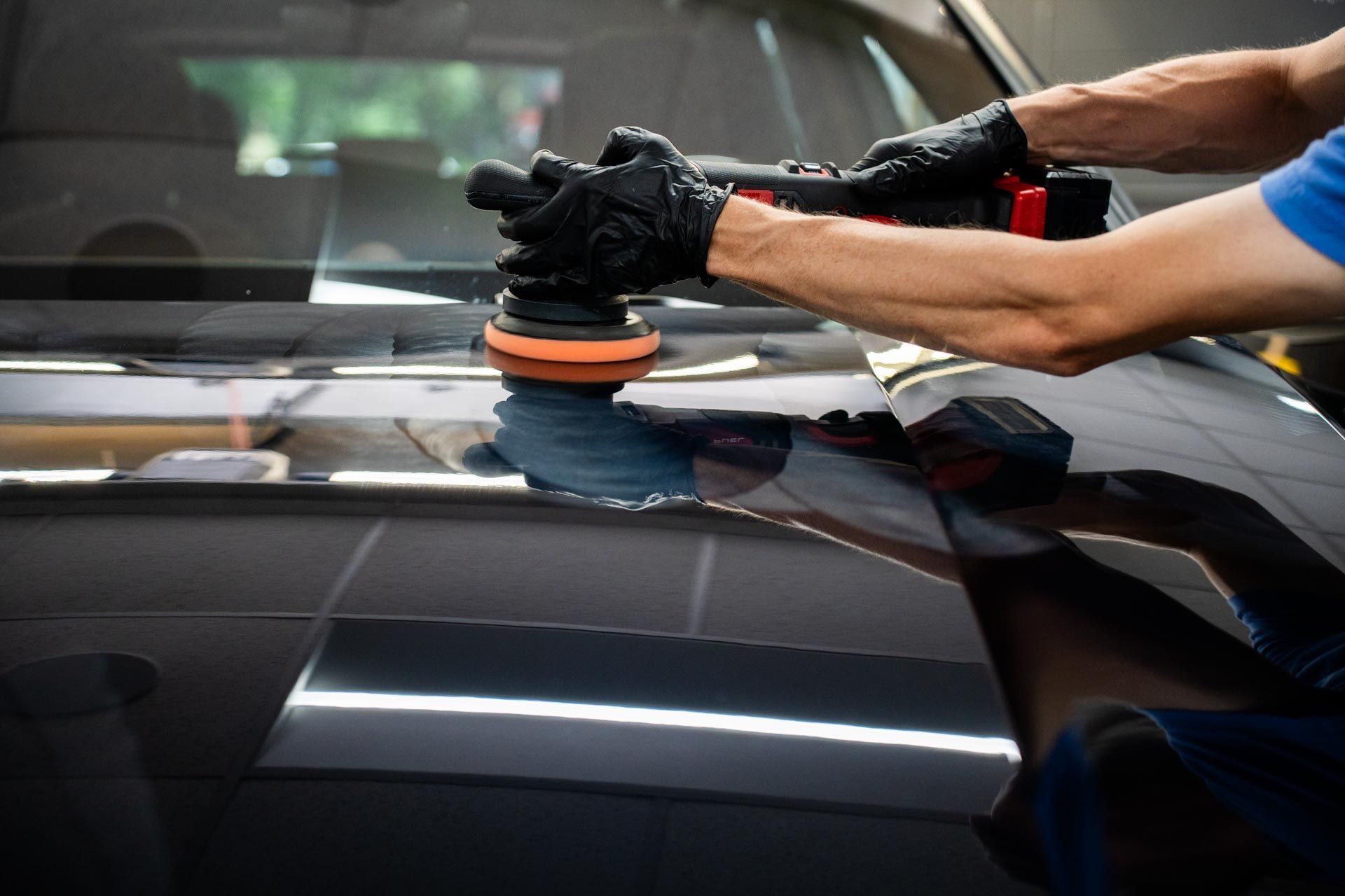 Person polishing a black car with an electric buffer, wearing black gloves.