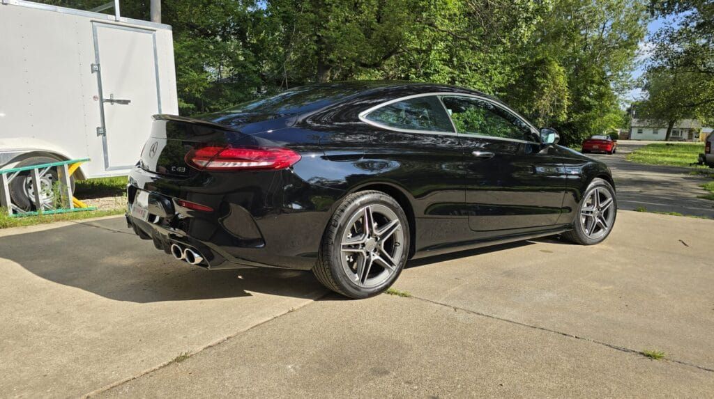 Black Mercedes-Benz coupe parked on a paved driveway, in front of a white trailer, trees, and a house.