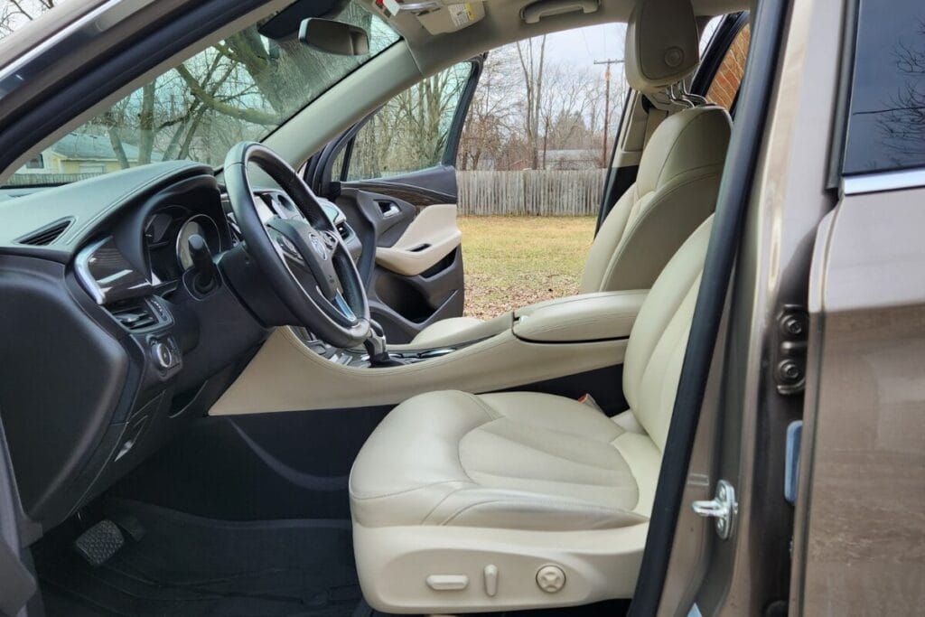 Tan leather car interior; driver's seat, dashboard, steering wheel visible. Door open, revealing outdoor setting.