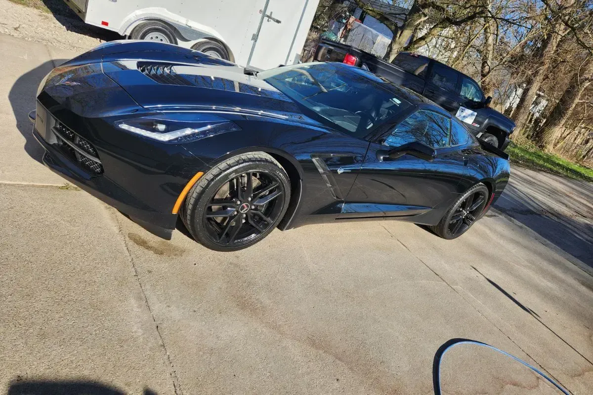 Black Corvette sports car parked on a driveway, beside a white trailer and a black truck.