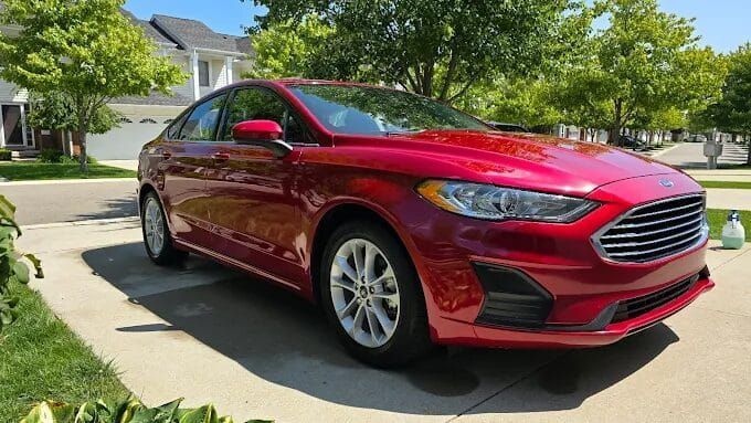 Red Ford Fusion sedan parked on a driveway in front of a house on a sunny day.