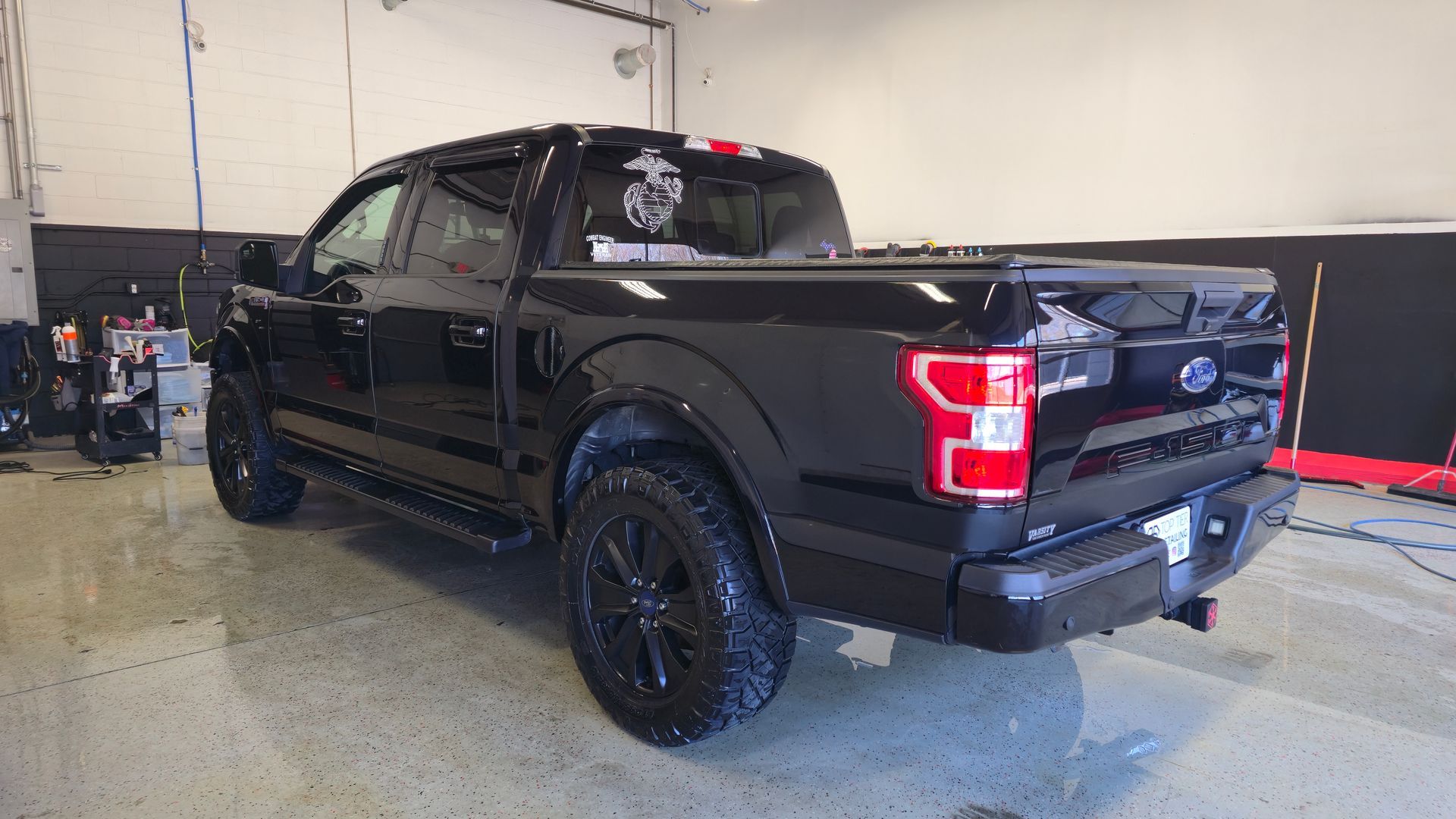 Black Ford F-150 pickup truck in a garage. It's been washed, shiny, black rims, and a tonneau cover.