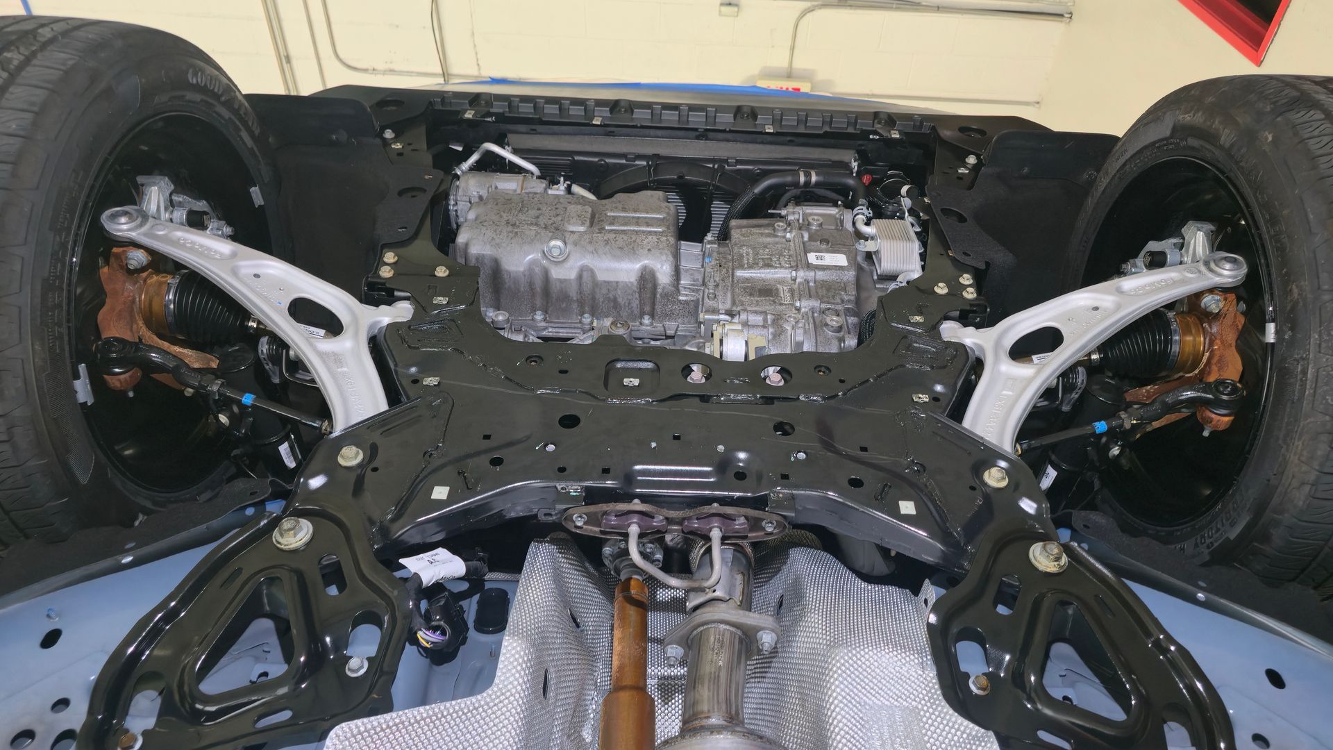 Undercarriage of a vehicle, showing suspension, engine, and tires. Black frame, silver engine, and gray underbody panels.