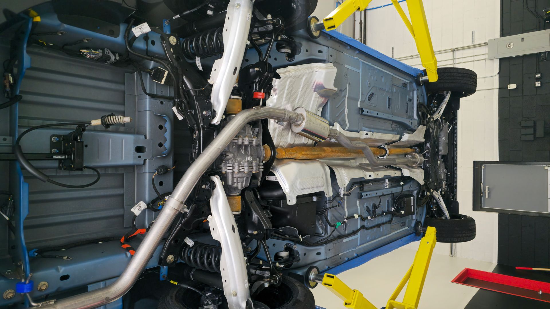 Underside of a blue car lifted on yellow jacks in a garage. Exhaust system visible.