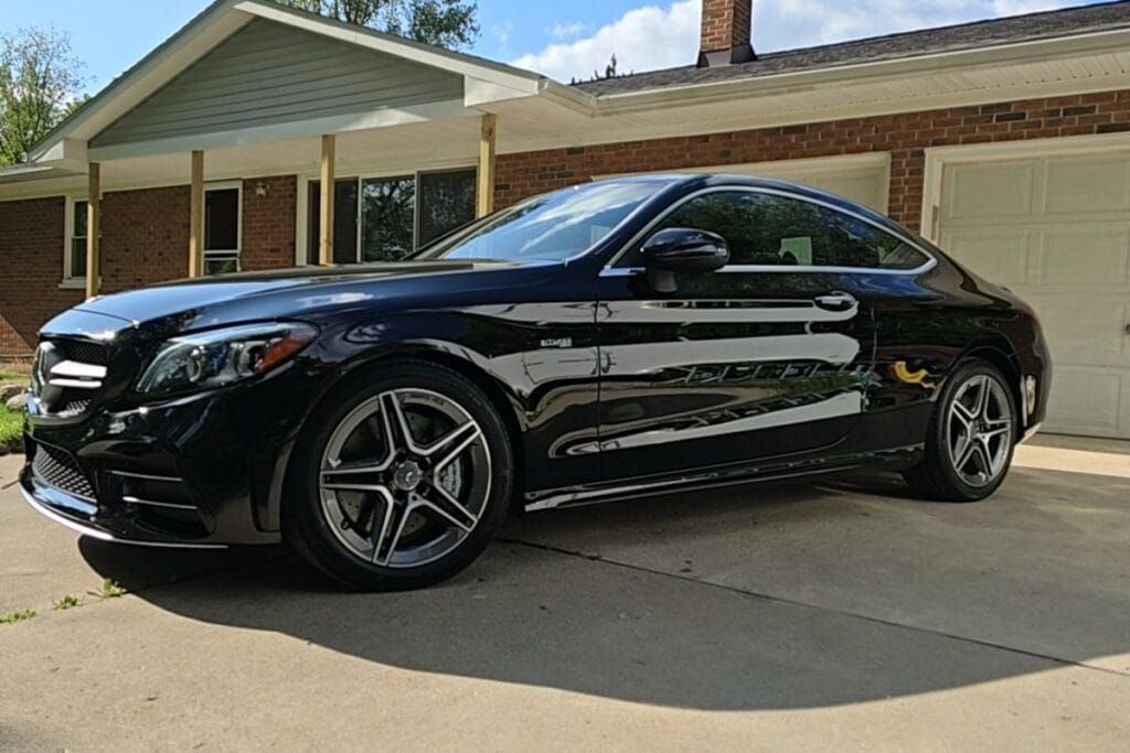 Black Mercedes coupe parked in front of a brick house with a two-car garage on a sunny day.