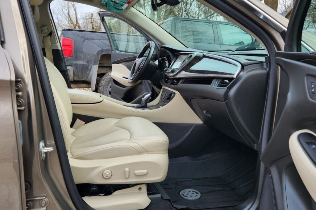 Beige car interior with opened driver's side door, white leather seats, black dashboard and floor mats.