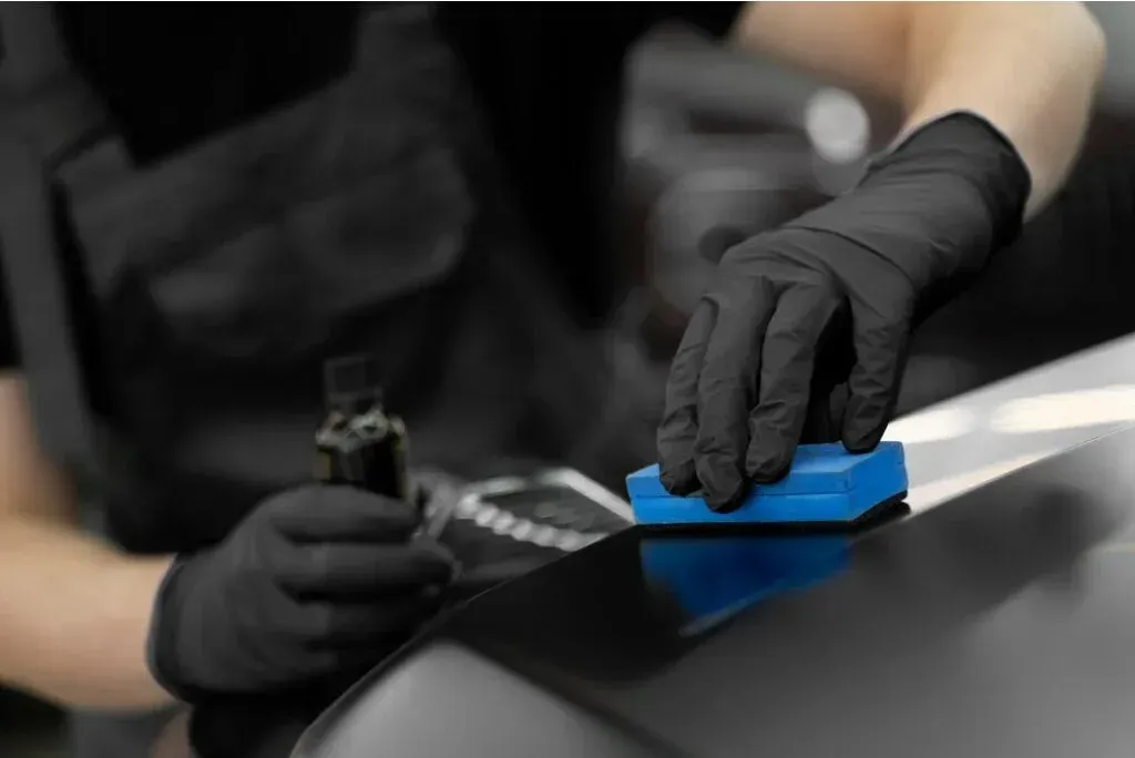 Person wearing black gloves applying blue sealant to a black car surface.