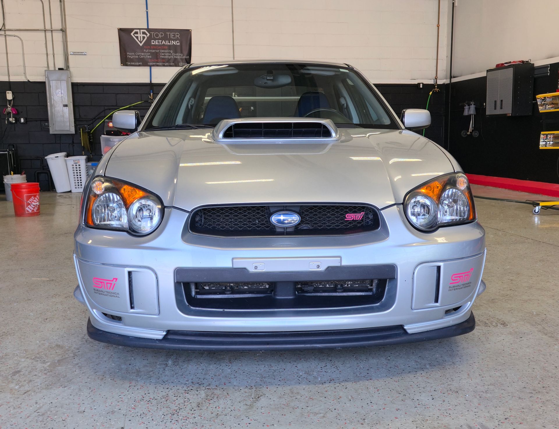 Silver Subaru WRX sedan parked indoors, front view. Black grille, hood scoop, pink accents.