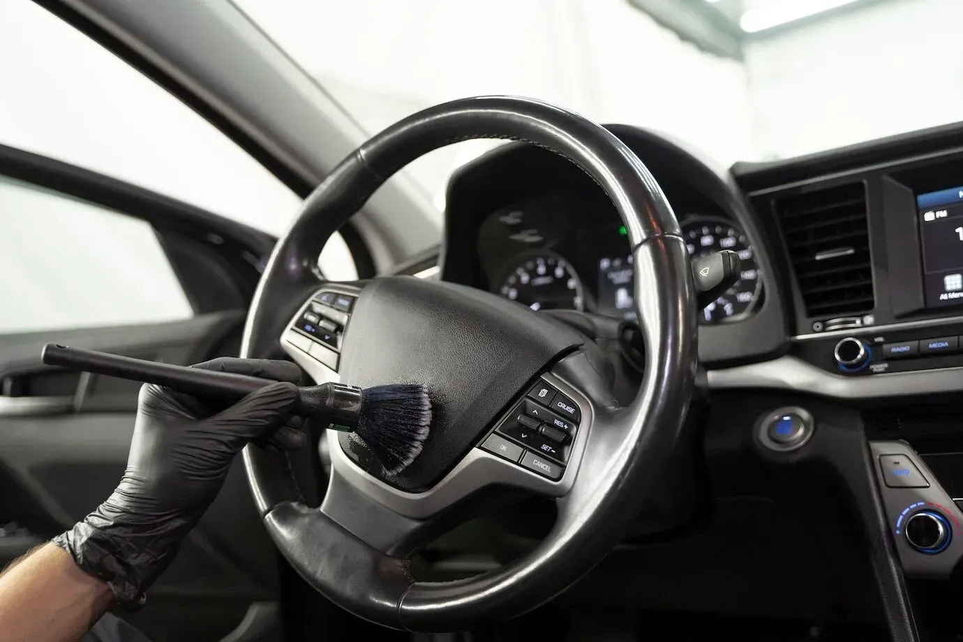 A gloved hand cleans a car steering wheel with a brush.
